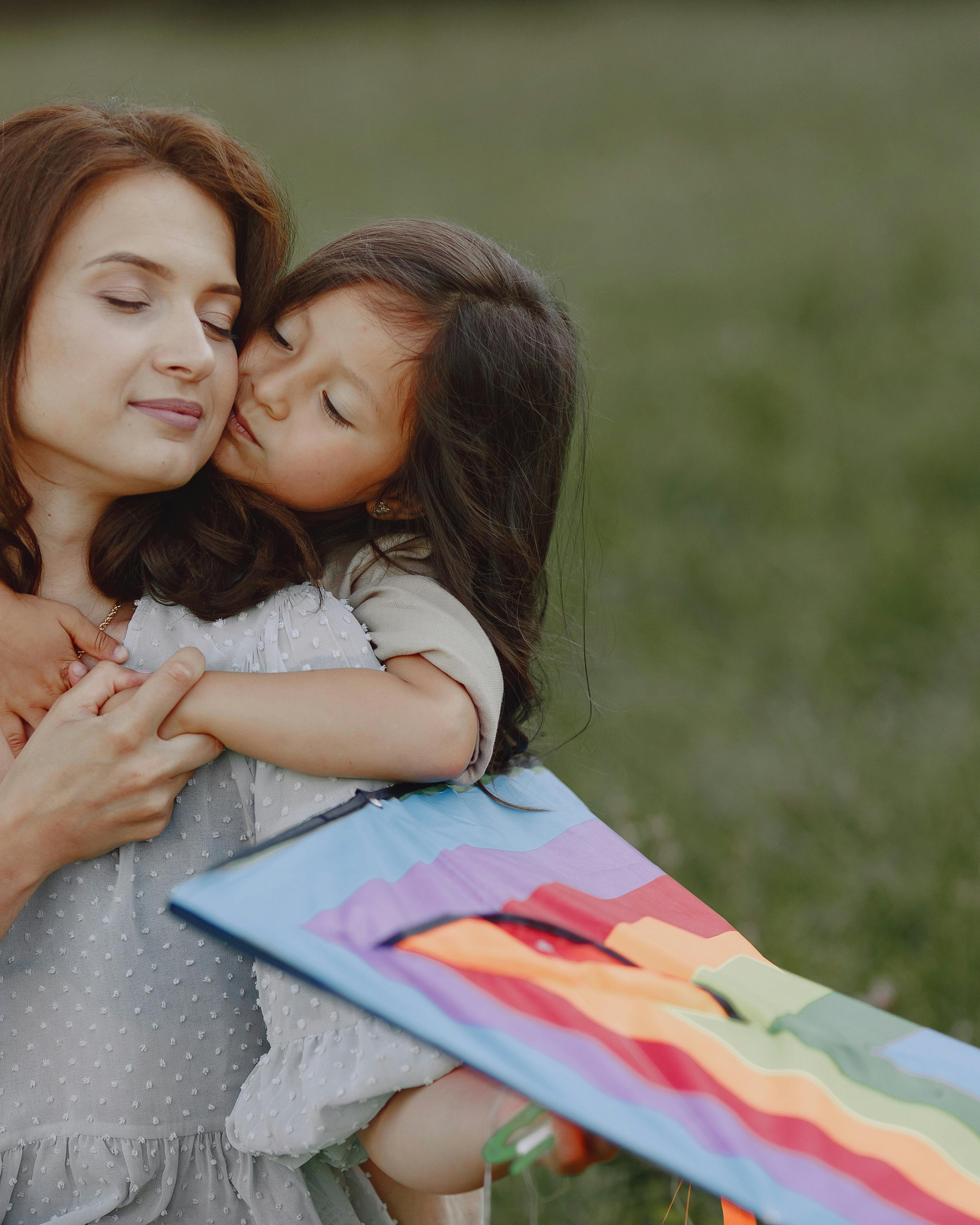 Daughter Kissing Mother With Kite In Meadow