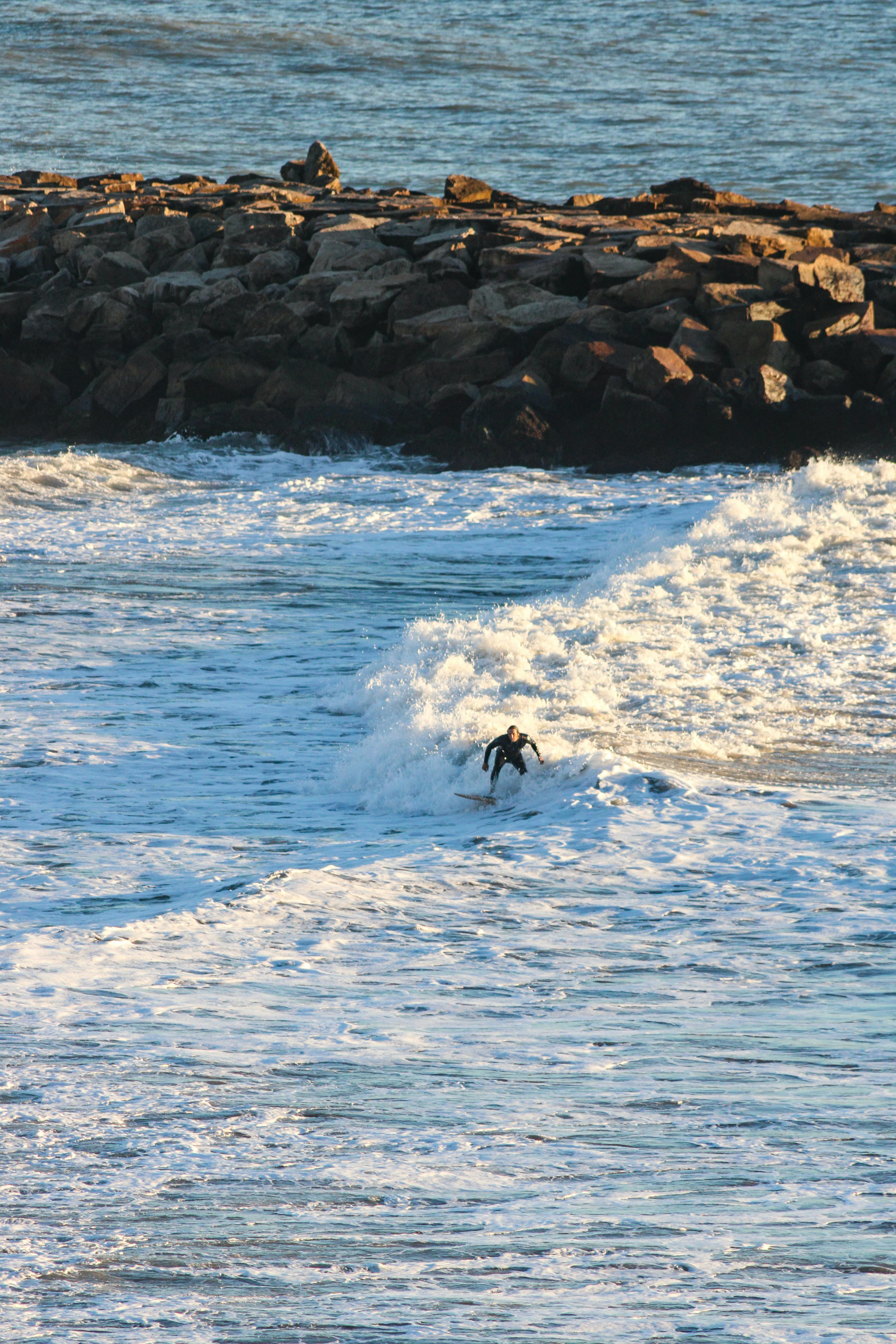 Surfer Riding Wave Near Rocky Breakwater
