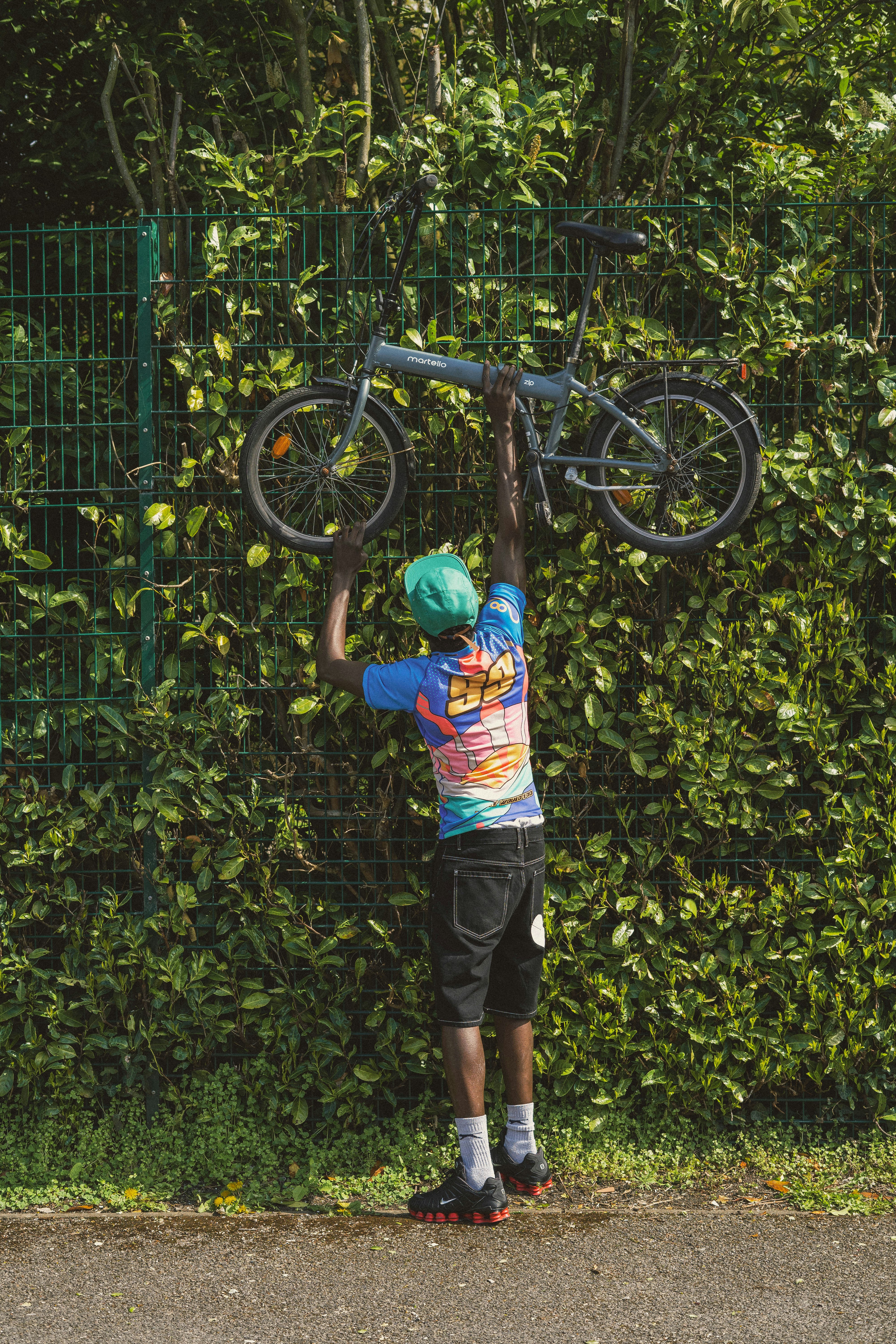 Man Lifting Folding Bike Against Green Fence